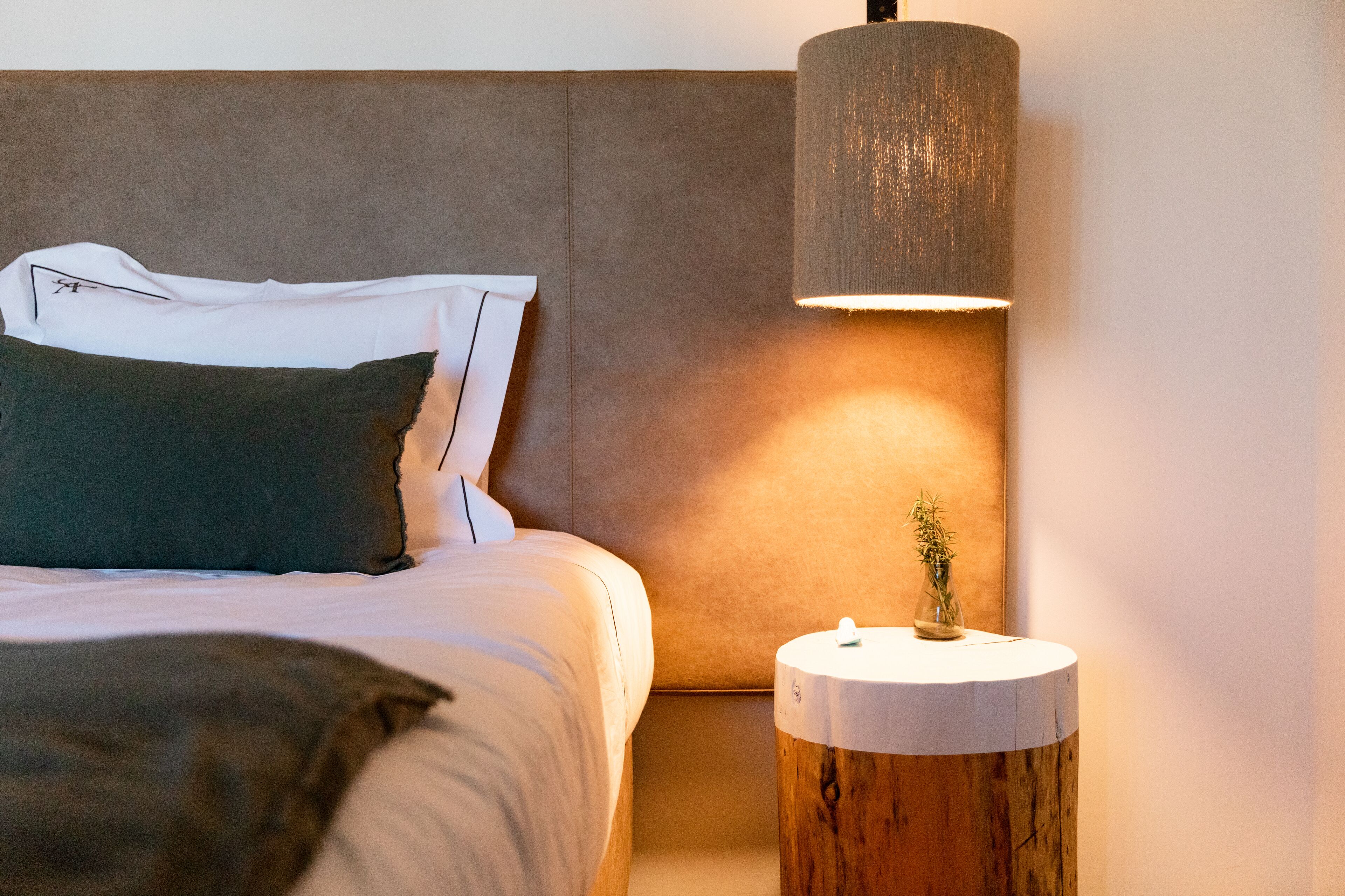 Una cama bien hecha con almohadas, una lámpara de noche y una mesita de madera con un florero.