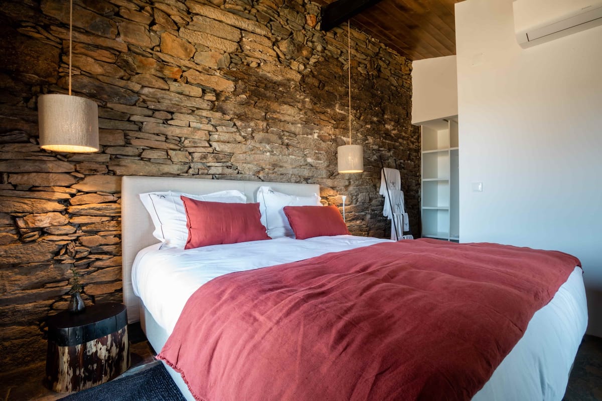 Een slaapkamer met een stenen muur, een groot bed met rode beddengoed en twee hanglampen.