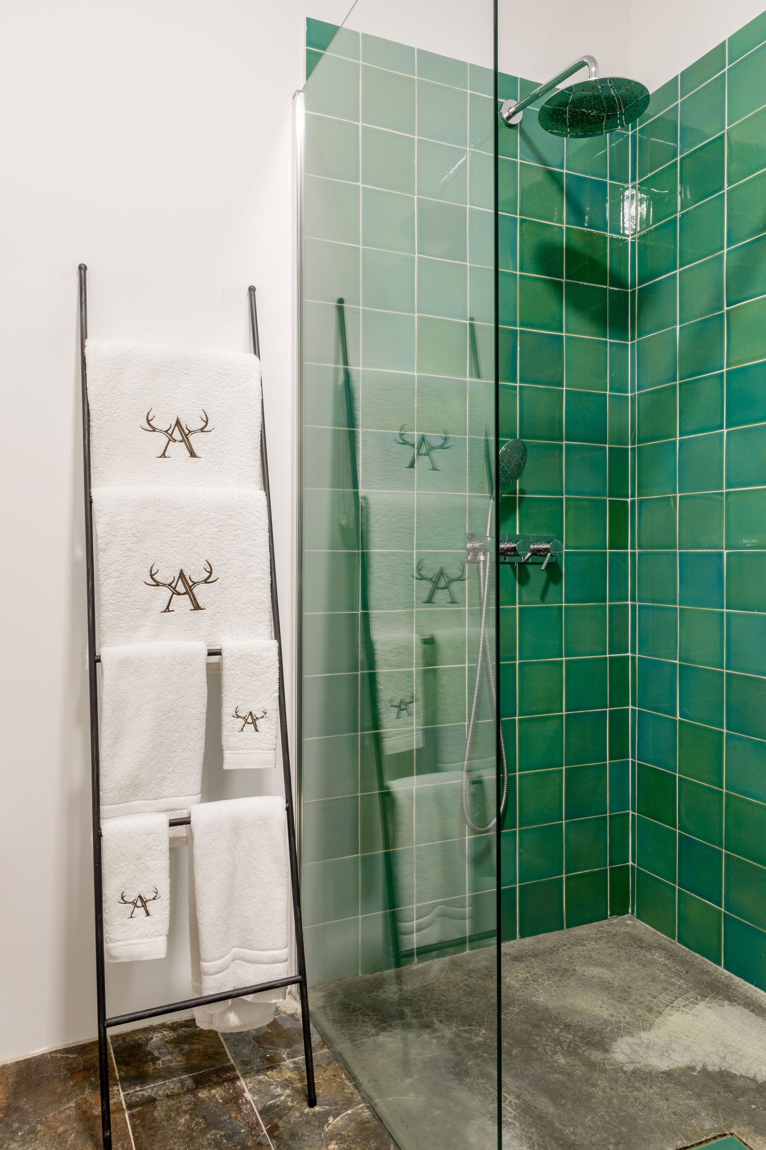 Ducha con mampara de vidrio, pared con azulejos verdes, un perchero con toallas blancas y un cabezal de ducha.