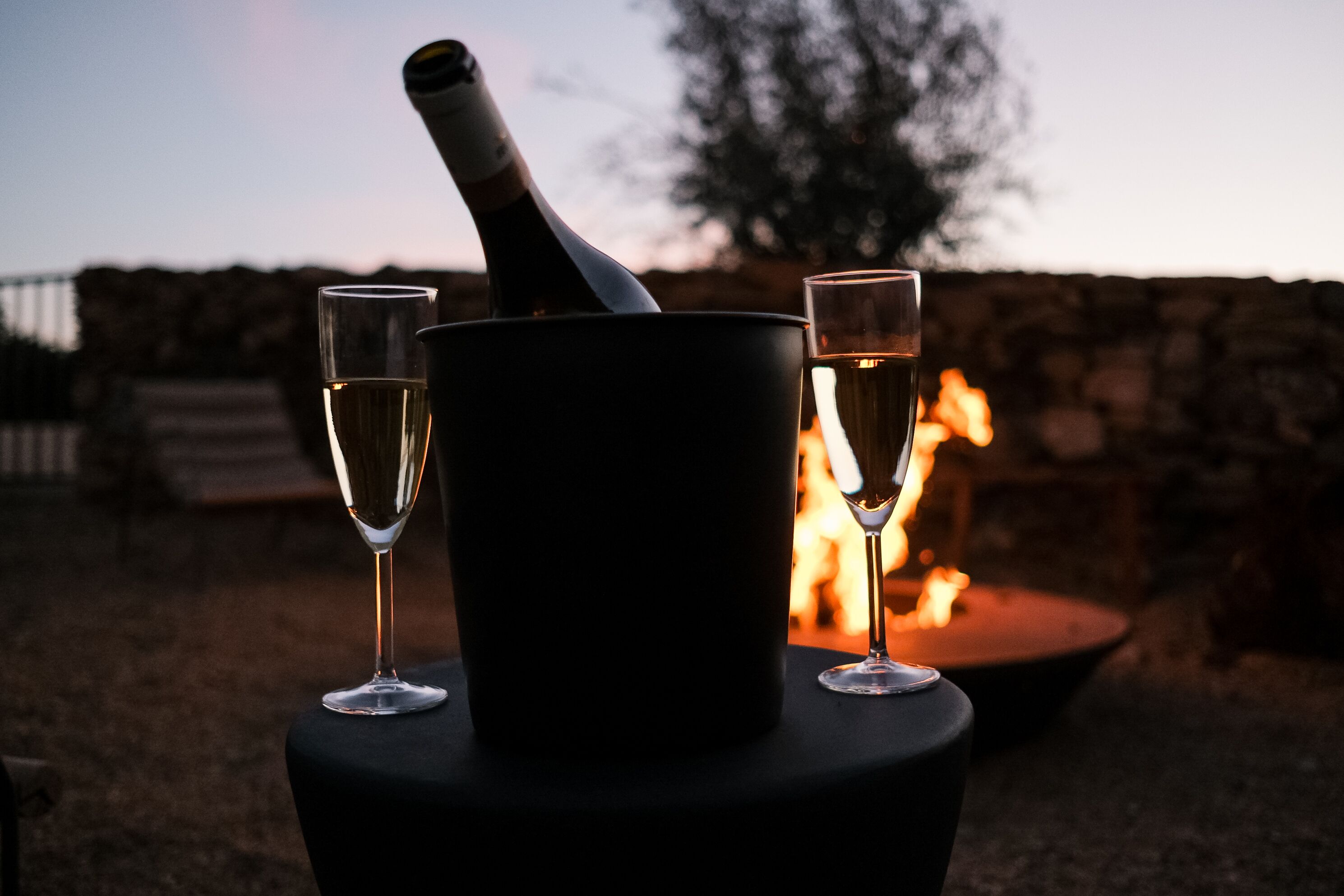 A bottle of champagne and two glasses on a round table with a fire in the background.