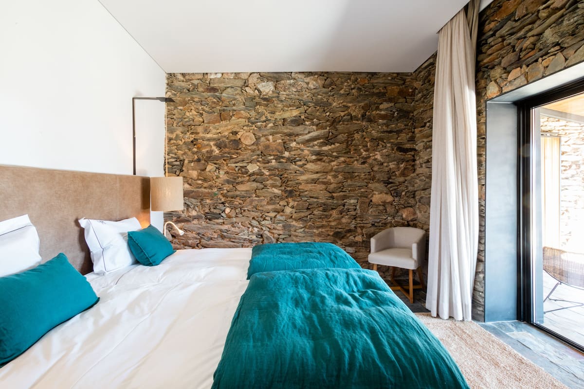 Een slaapkamer met een stenen muur, een bed met wit en blauwgroen beddengoed, een bedlampje en een stoel bij een raam.