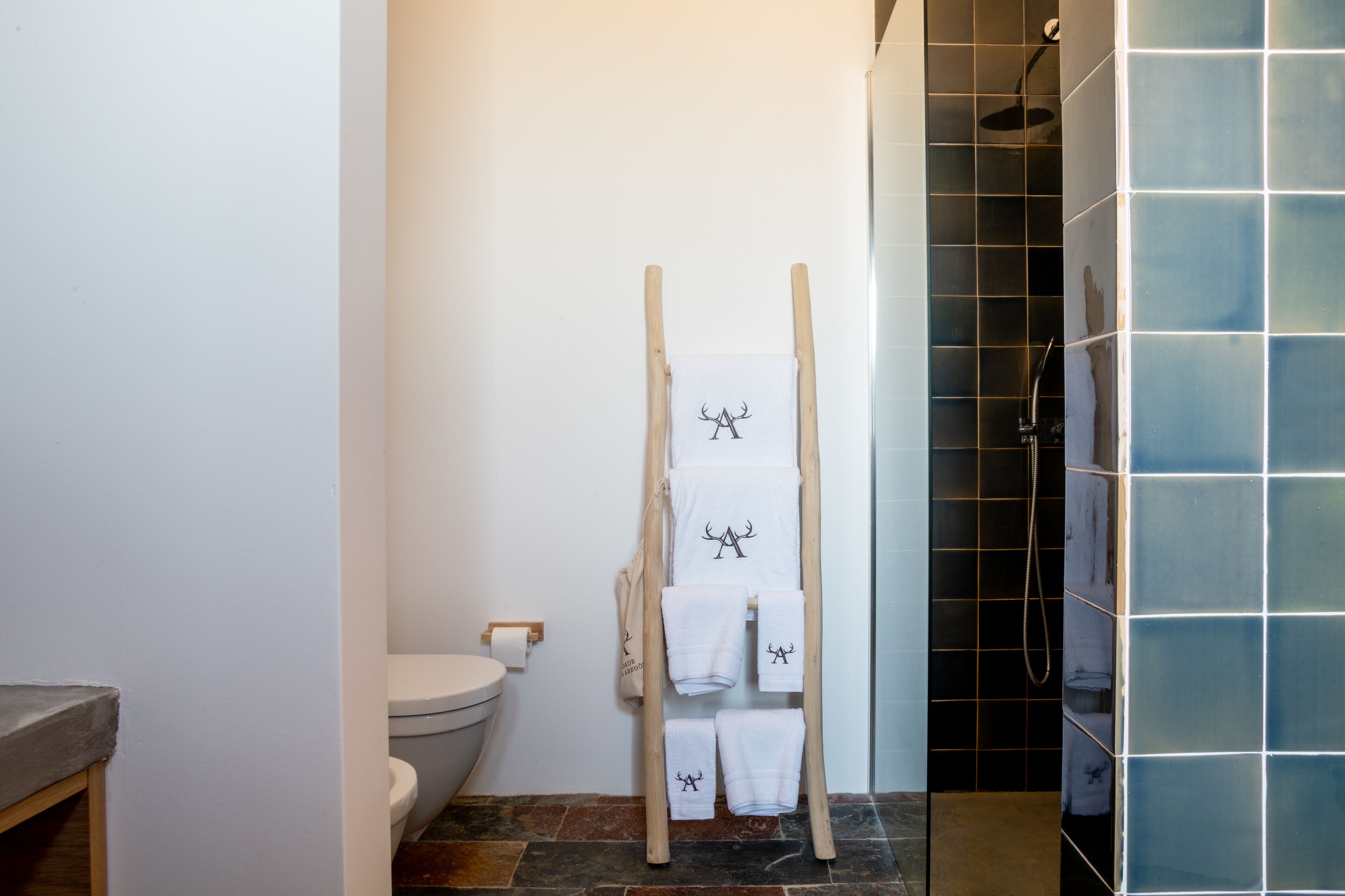 Een badkamer met een houten handdoekrek vol witte handdoeken, een toilet en een douchegedeelte met blauwe tegels.
