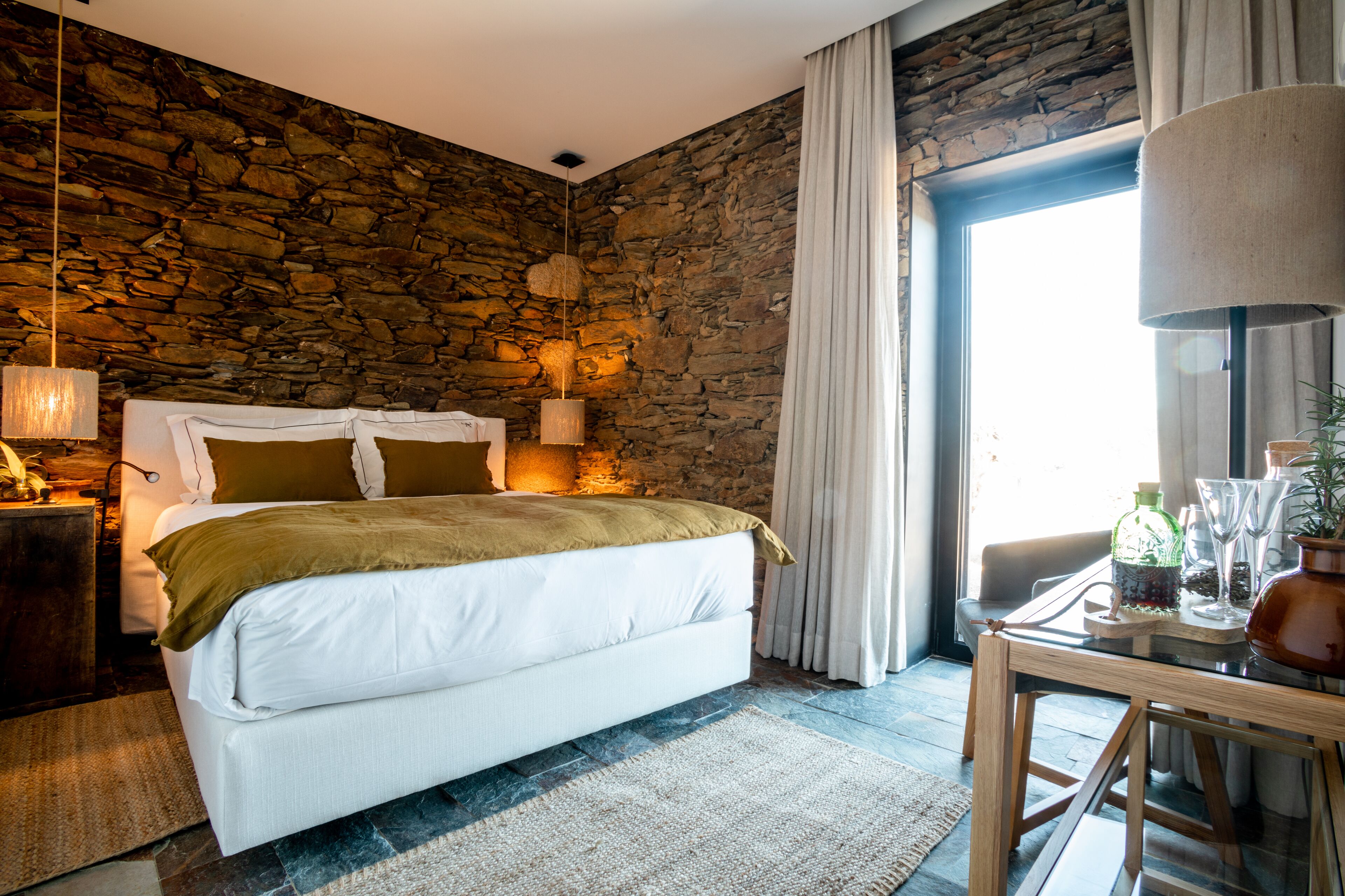 Un dormitorio con pared de piedra, cama con ropa blanca, mesa de madera con un jarrón y una lámpara colgante.