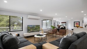 A modern living room with a sofa set, a coffee table, and a TV. There are large windows with blinds, a kitchen area in the background, and a dining table with chairs. - Hoddle Lane – rural views, fireplace and air conditioning (Robertson)