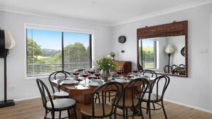 A dining room with a wooden table set for six, surrounded by six chairs. There is a vase with flowers on the table, and a clock on the wall.