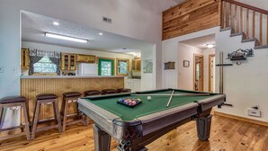 Een pooltafel in een ruimte met houten vloeren, een bar met krukjes en een trap.