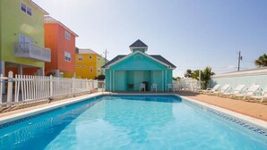 Una piscina con un edificio turquesa al fondo, rodeada de edificios coloridos y una valla blanca.
