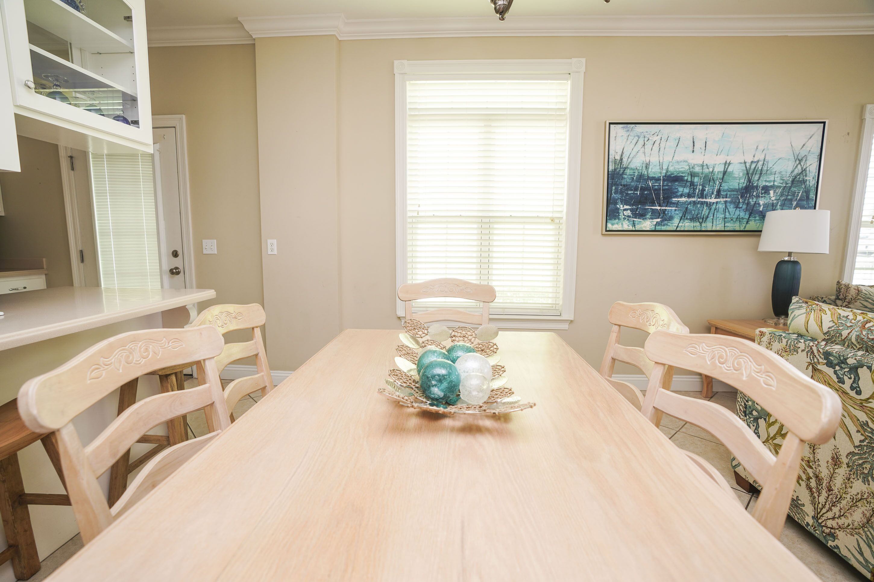Uma área de refeições com mesa e cadeiras de madeira, um centro de mesa decorativo de vidro, uma janela com persianas e um quadro na parede.