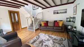A living room with a brown leather sofa, a grey patterned rug, wooden flooring, a staircase with white railings, and a television on a black stand. - Charming Barn Conversion on Private Estate (Kilmarnock)