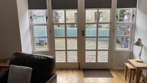 A room with a black leather sofa, a wooden table with a lamp, and a view through a set of French doors to a courtyard with a stone floor, a blue fence, and a parked car. - Charming Barn Conversion on Private Estate (Kilmarnock)