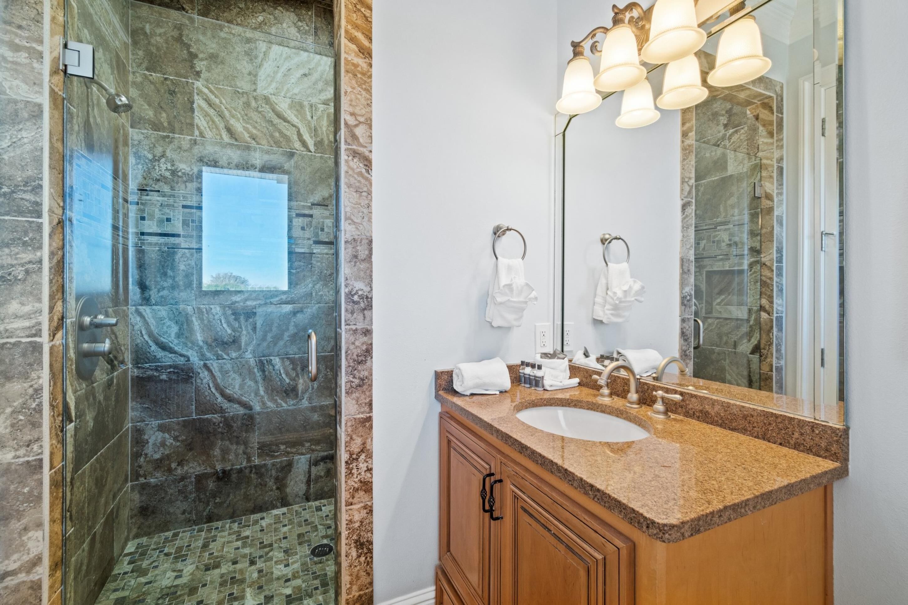 A bathroom with a glass-enclosed shower, a granite countertop sink, and a large mirror with multiple light fixtures.