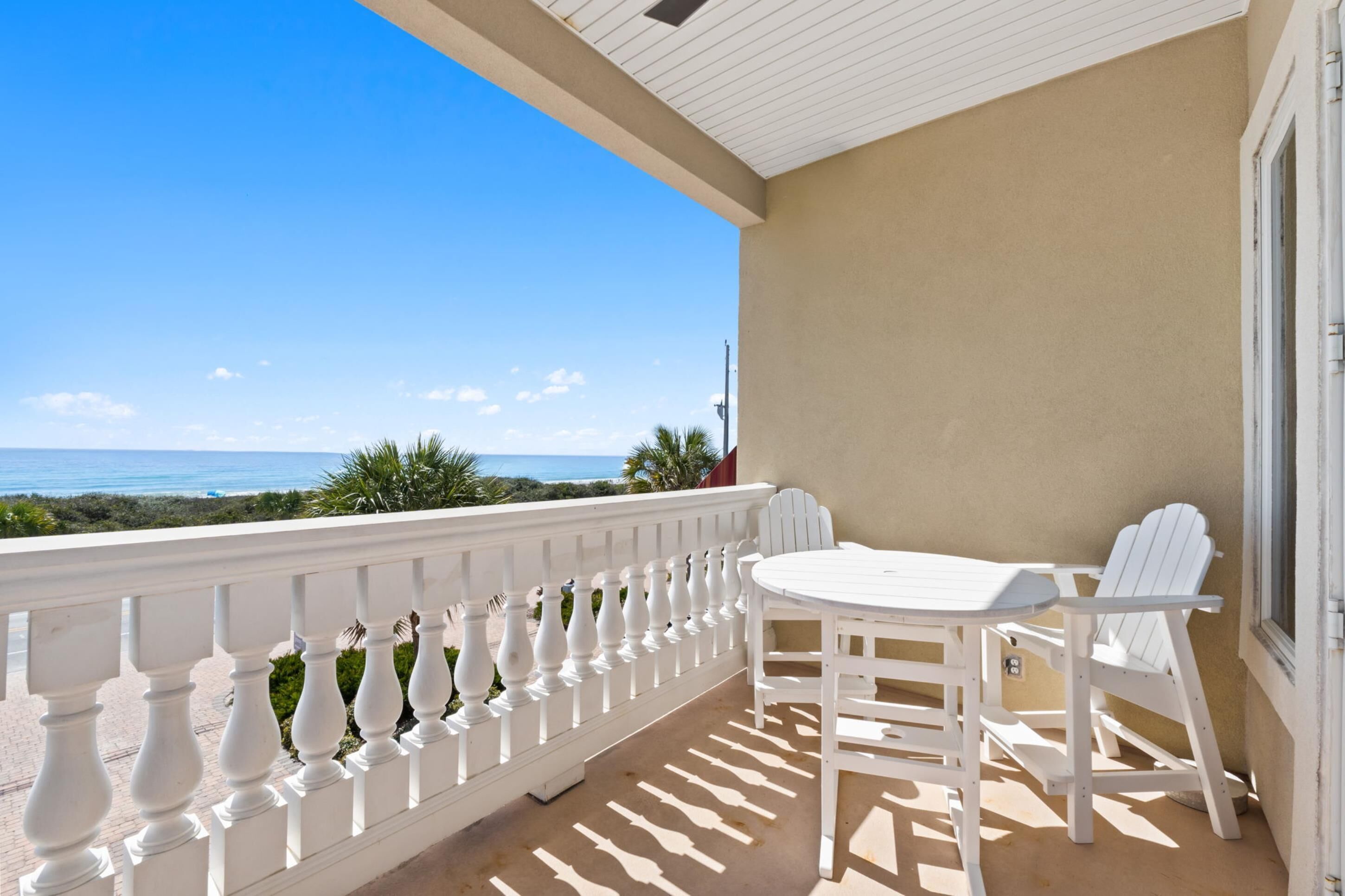 A balcony with a white railing, a table, and two chairs, overlooking a beach.