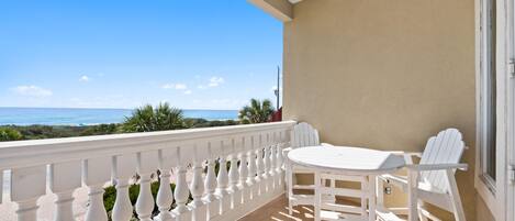 A balcony with a white railing, a table, and two chairs, overlooking a beach.
