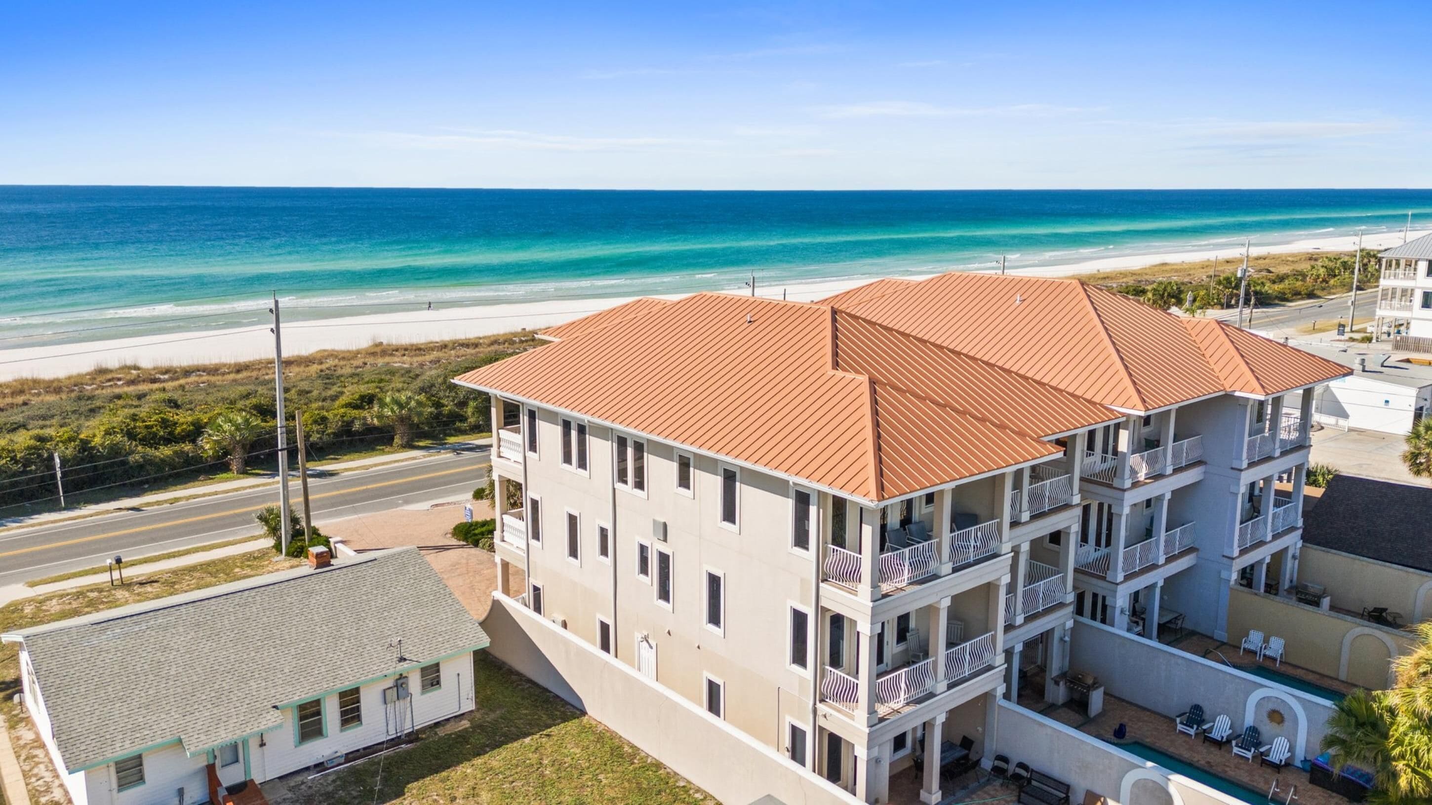 A multi-story hotel with a red-tiled roof, a pool area, and a view of the beach and ocean.
