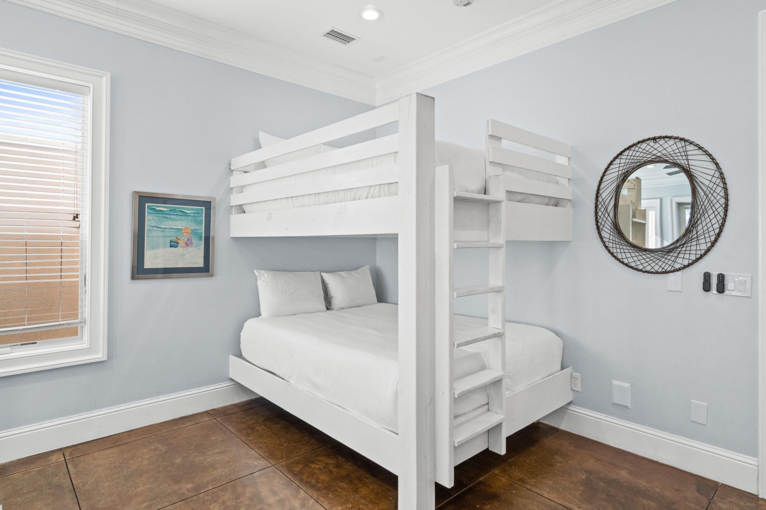 A room with a bunk bed, a framed picture on the wall, a round mirror, and a window with blinds.