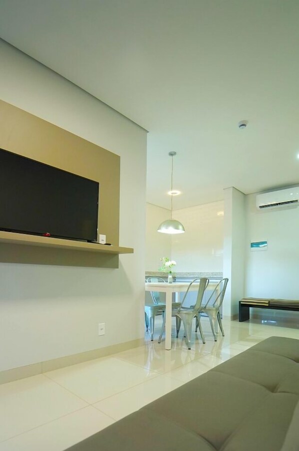 A modern living room with a flat-screen TV mounted on a wall, a dining area with a table and chairs, and a sofa.