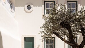 Une cour avec un bâtiment blanc, une porte verte et un grand arbre. Des tables et des chaises y sont disposées pour permettre de prendre les repas en plein air.