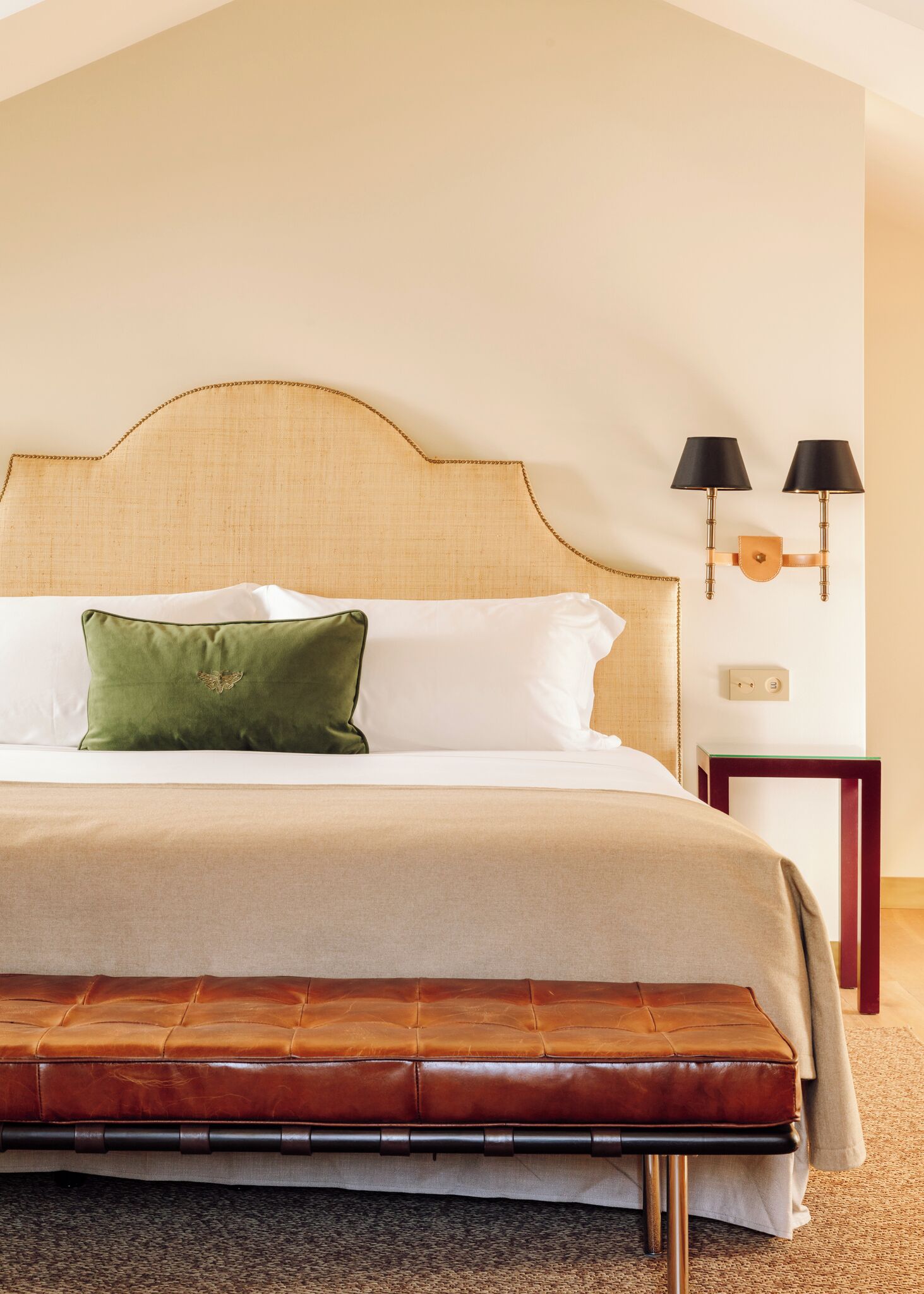 A neatly made bed with a green pillow, a wooden bench, and two wall-mounted lamps.