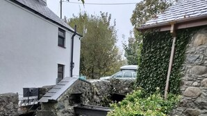 Un pont de pierre enjambe un petit ruisseau où l’eau forme une cascade, le tout étant entouré de verdure et bordé d’un mur de pierres.