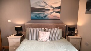 Une chambre avec un grand lit, deux tables de chevet avec des lampes, une commode, un tableau représentant un paysage de montagne accroché au mur, et une fenêtre avec des rideaux.