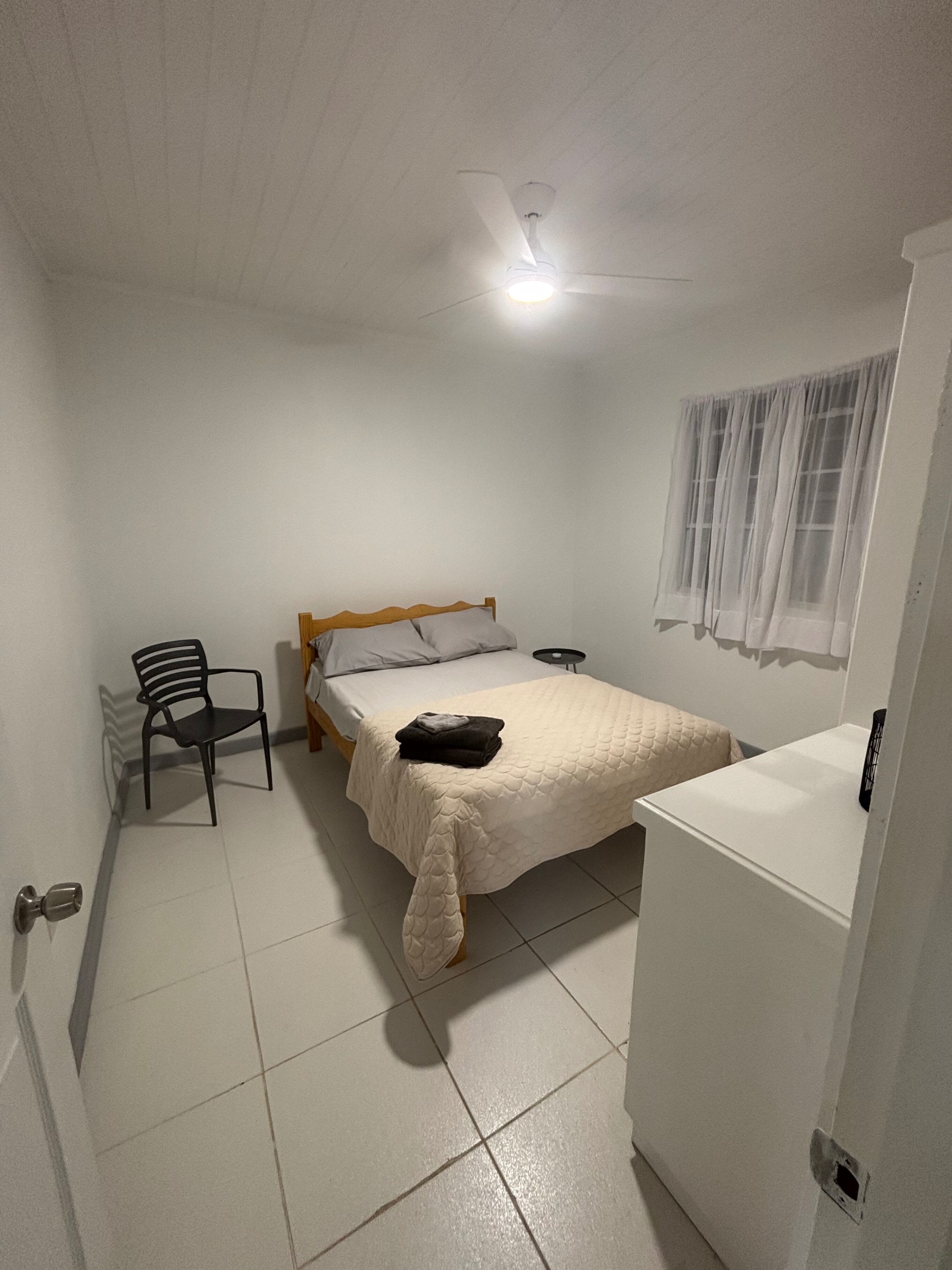 A small, clean room with a single bed, a chair, a small table, and a window with curtains.