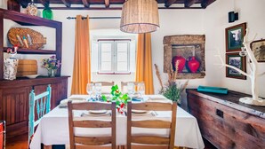 Un comedor con una mesa de madera dispuesta para cuatro personas, una lámpara colgante de fibras tejidas y una rústica pared de piedra con jarrones decorativos.