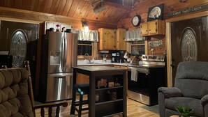 Ein Blockhaus-Interieur mit Holzwänden, einem Deckenventilator, einer Küchenzeile mit Geräte und einer Sitzecke mit Sessel und Sofa.