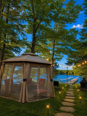 A gazebo with a covered seating area, surrounded by trees and a pathway leading to a dock.