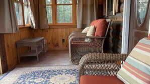 Ein gemütliches Blockhaus-Interieur mit Korbmöbeln, einem gemusterten Teppich und großen Fenstern mit Blick auf den Wald.