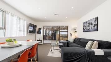 A modern living room with a black sofa, orange chairs, a dining table, and a TV mounted on the wall.