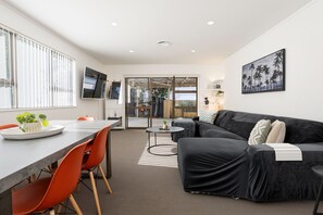 A modern living room with a black sofa, orange chairs, a dining table, and a TV mounted on the wall.