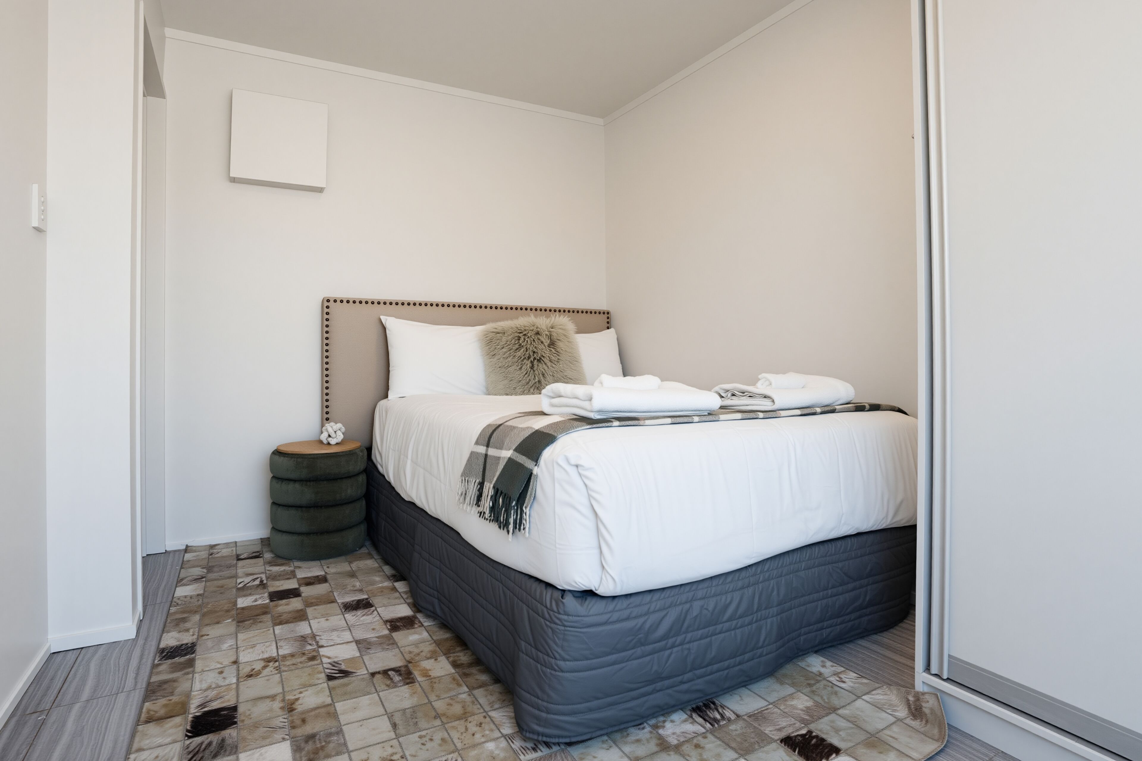 A bedroom with a bed, a bedside table, a small stool, and a checkered floor.