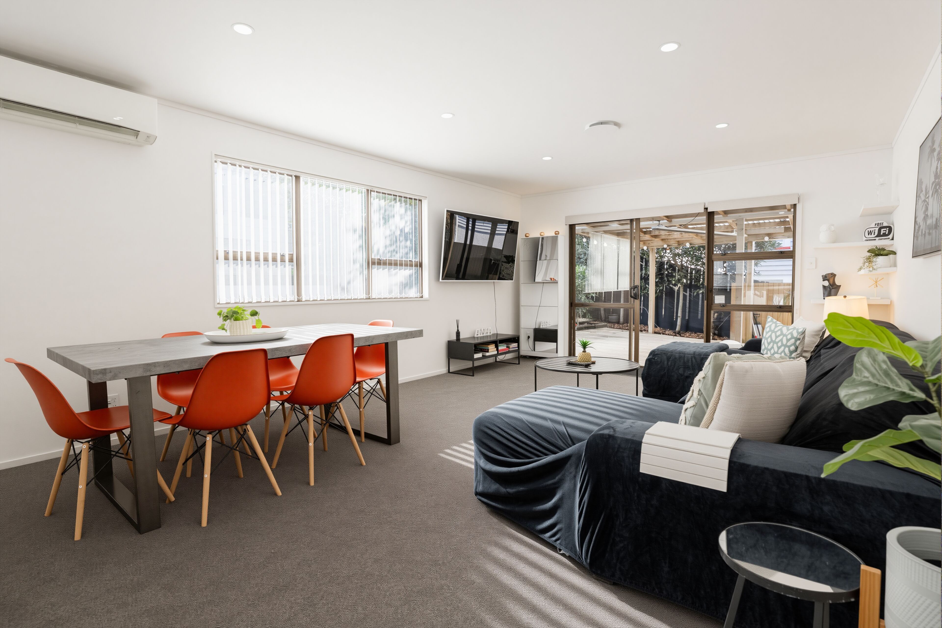 A modern living space with a dining area, a sofa, a TV, and a sliding glass door leading to an outdoor area.