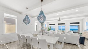 A spacious dining area with a white table and chairs, a chandelier, and a view of the ocean through large windows. - Wilder Dreams - Gulf Front, Pool, Gated Community, Steps to Rosemary Beach (Seacrest)