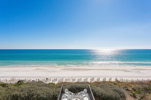 Blick auf einen Strand, mit blauem Himmel, sandigem Ufer und einem eingezäunten Bereich mit Stühlen.