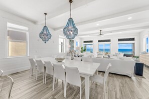 Ein geräumiger Essbereich mit einem weißen Tisch und Stühlen, einer Chandelier und Meerblick durch große Fenster.