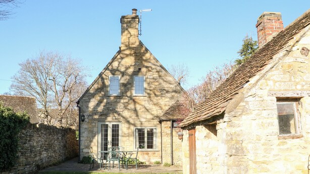 A stone house with a sloped roof, a chimney, and a small garden with a table and chairs.