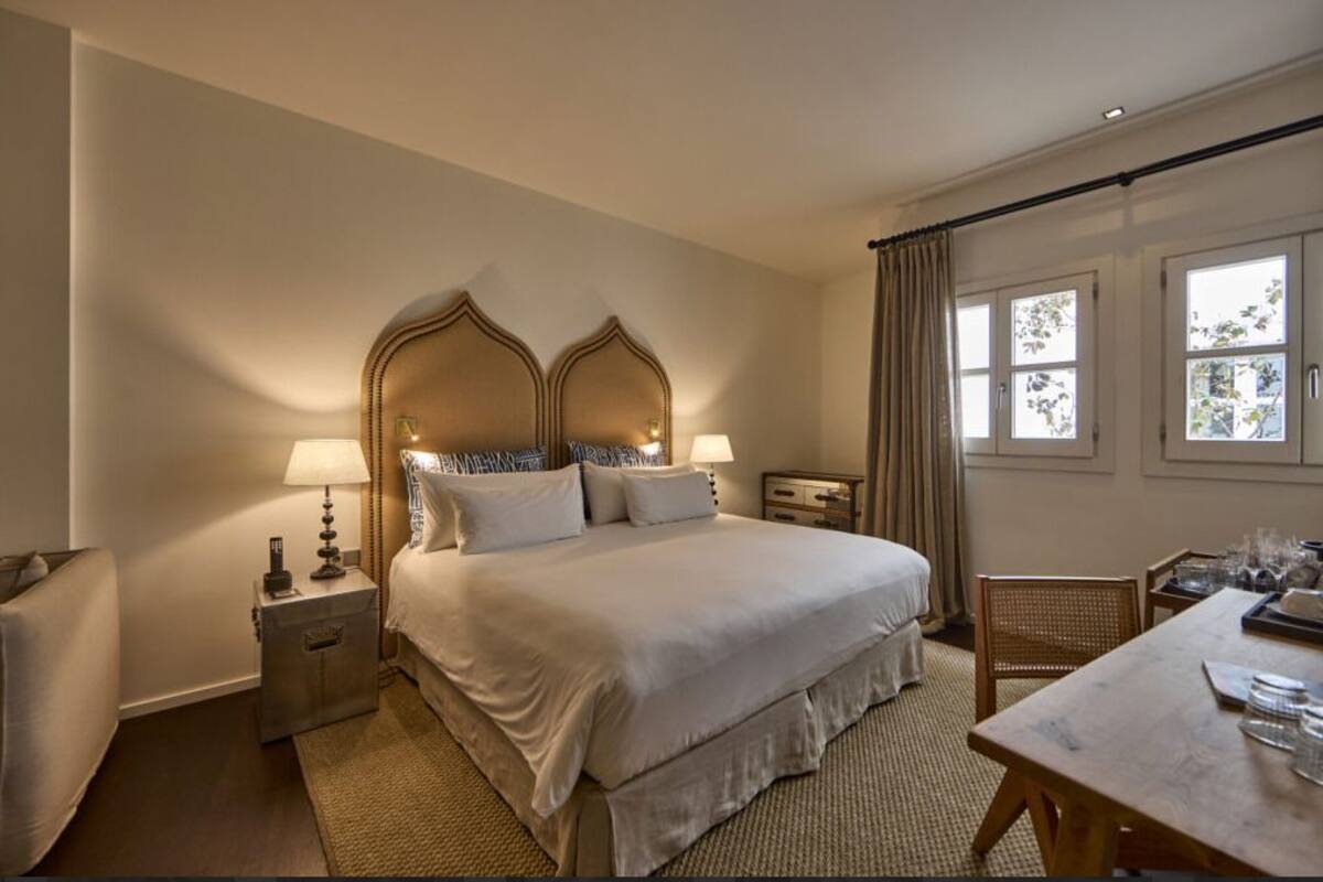 Een hotelkamer met een groot bed, twee nachtkastjes met lampen, een houten stoel en een eettafel gedekt voor twee personen.