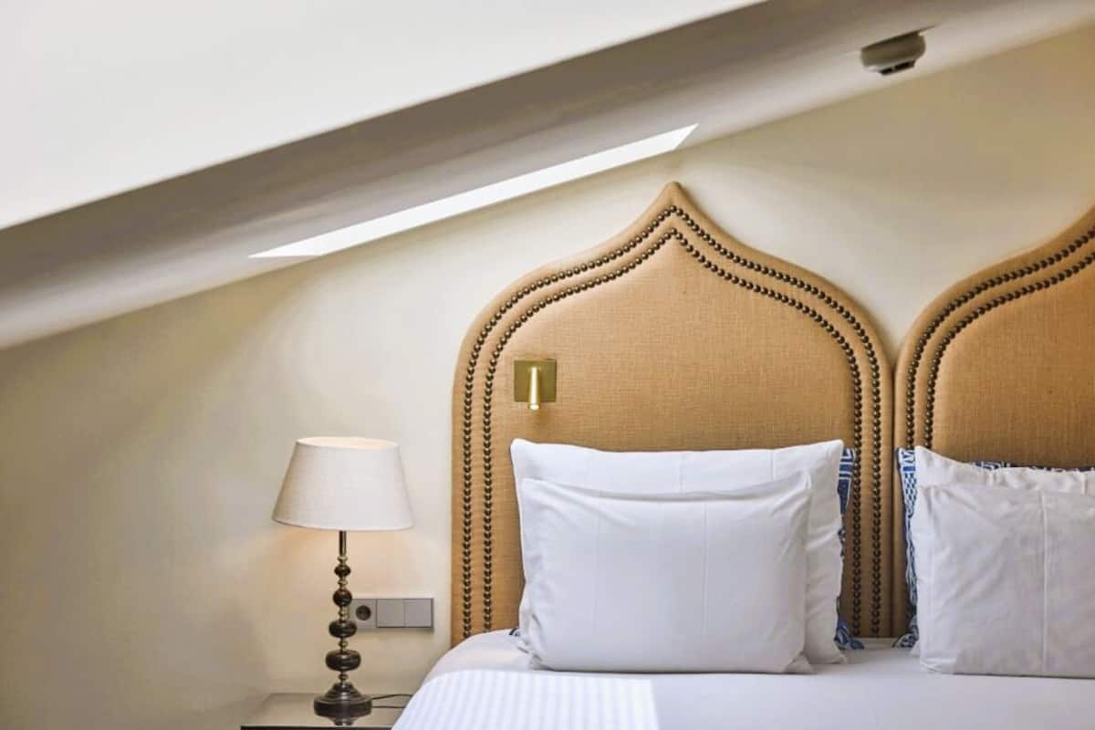 Een hotelkamer met een hoog, decoratief hoofdeinde, een bedlampje en een net opgemaakt bed met witte lakens.
