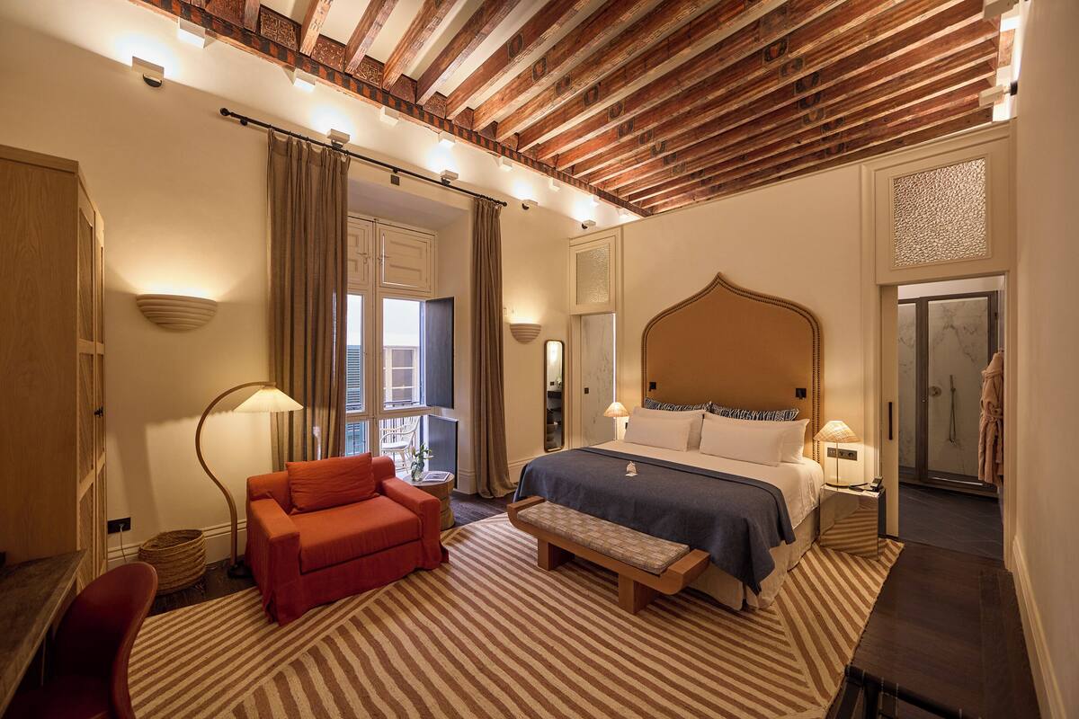 Een slaapkamer met een groot bed, een bankje, een rode stoel, een vloerlamp en een houten plafond.