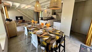 A dining area with a long table set for six, two hanging wicker lamps, a staircase in the background, and a kitchen area visible through an open door.