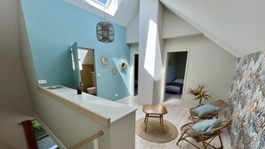 A spacious loft with a high ceiling, a large skylight, a wooden staircase, a dining area with wicker chairs, and a bedroom with a bed and a large window.