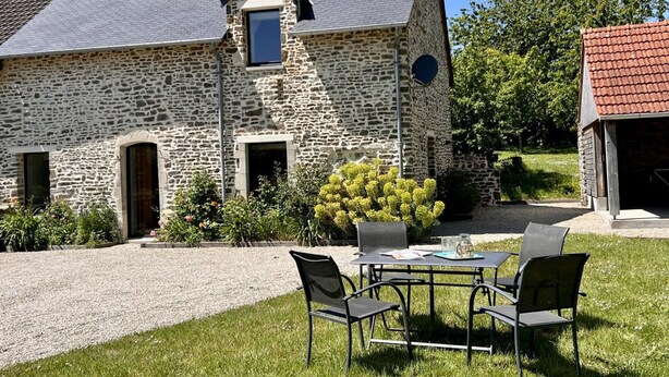 A stone house with a sloped roof, a chimney, and a small window. There is a gravel driveway leading to a table with chairs in the front yard.