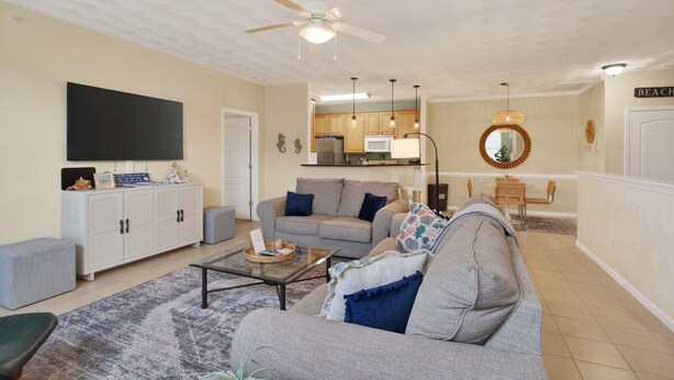 A living room with a sofa, coffee table, and TV. There's a kitchen in the background with wooden cabinets and a ceiling fan.