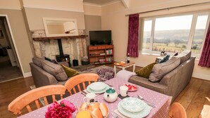 A cozy living room with a dining table set for a meal, a fireplace, a television, and a view of the countryside. - Healey Farm Cottage (Morpeth)
