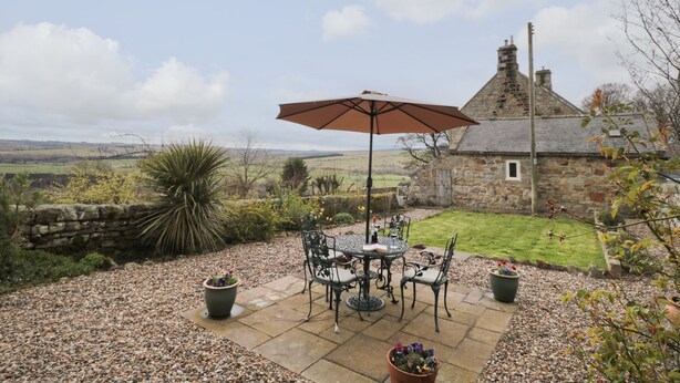 A stone house with a thatched roof, a patio with a table and chairs, and a view of the countryside. - Healey Farm Cottage (Morpeth)