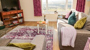 A living room with a sofa, a coffee table with a board game, a TV on a wooden stand, and a window with curtains. - Healey Farm Cottage (Morpeth)