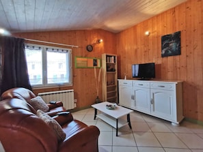 A cozy living room with a brown leather sofa, a white coffee table, a TV on a white cabinet, and a window with blinds. - Charming Retreat with Sauna and Terrace in Peaceful Hamaulet (Saint-Just-prés-Brioude)
