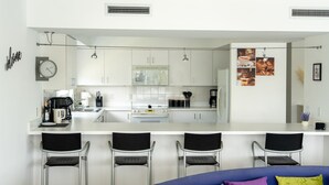 Uma cozinha moderna com armários brancos, uma ilha central, banquetas, uma máquina de café e um relógio.