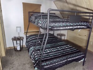 Een stapelbed met beddengoed met patroon, een klein tafeltje met een lamp en een raam met jaloezieën.